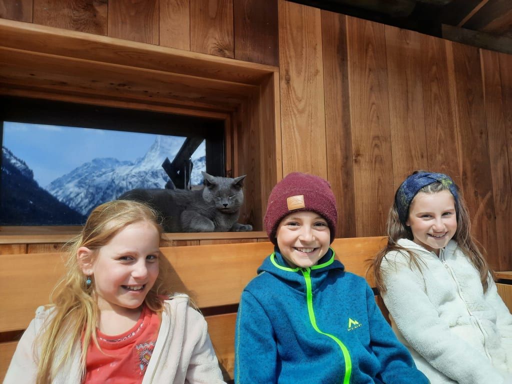 Drei Kinder und eine Katze auf einer Holzbank im Bauernhof, mit Bergblick durch ein Fenster im Hintergrund.