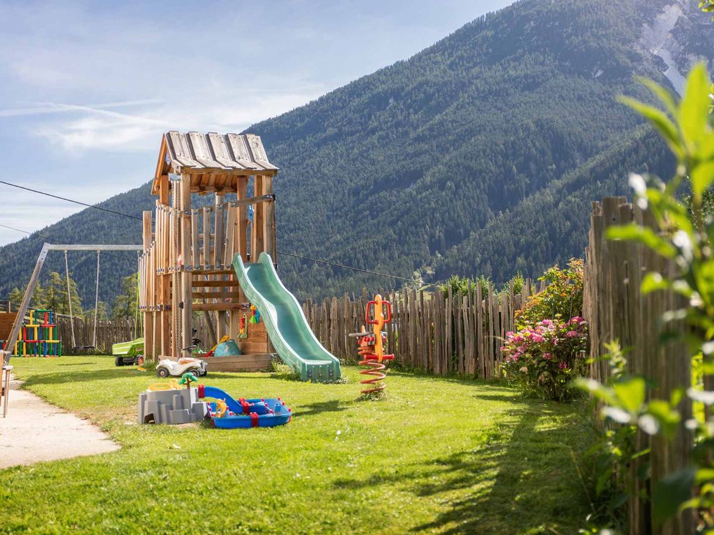 Kinderspielplatz auf dem Bauernhof mit Rutsche, Schaukel, Sandkasten und Federtier vor einer Bergkulisse.