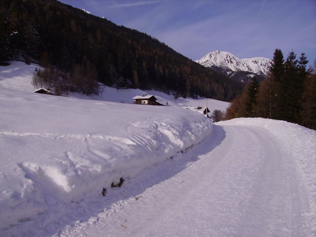 Ein verschneiter Weg führt durch die Winterlandschaft zum Bauernhof, umgeben von Wäldern und Bergen.