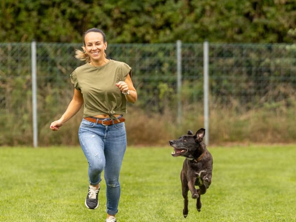 Eine Frau und ein Hund beim energiegeladenen Spiel auf der Rasenfläche des Bauernhofs.