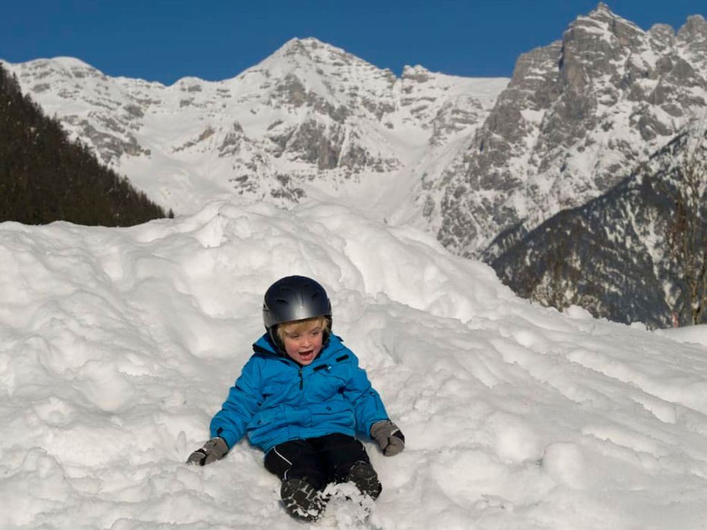 Ein Kind mit Helm rutscht im Schnee vor einer verschneiten Berglandschaft.