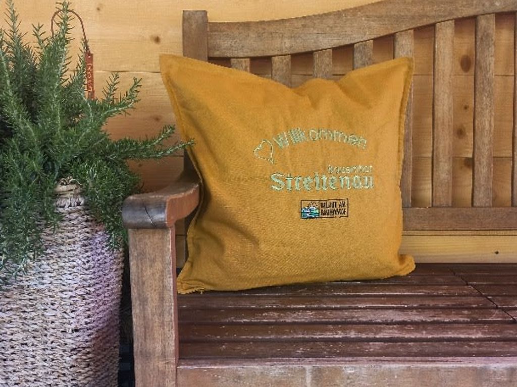 Ein gelbes Kissen mit der Aufschrift „Willkommen Bauernhof Streitenau“ auf einer Holzbank.
