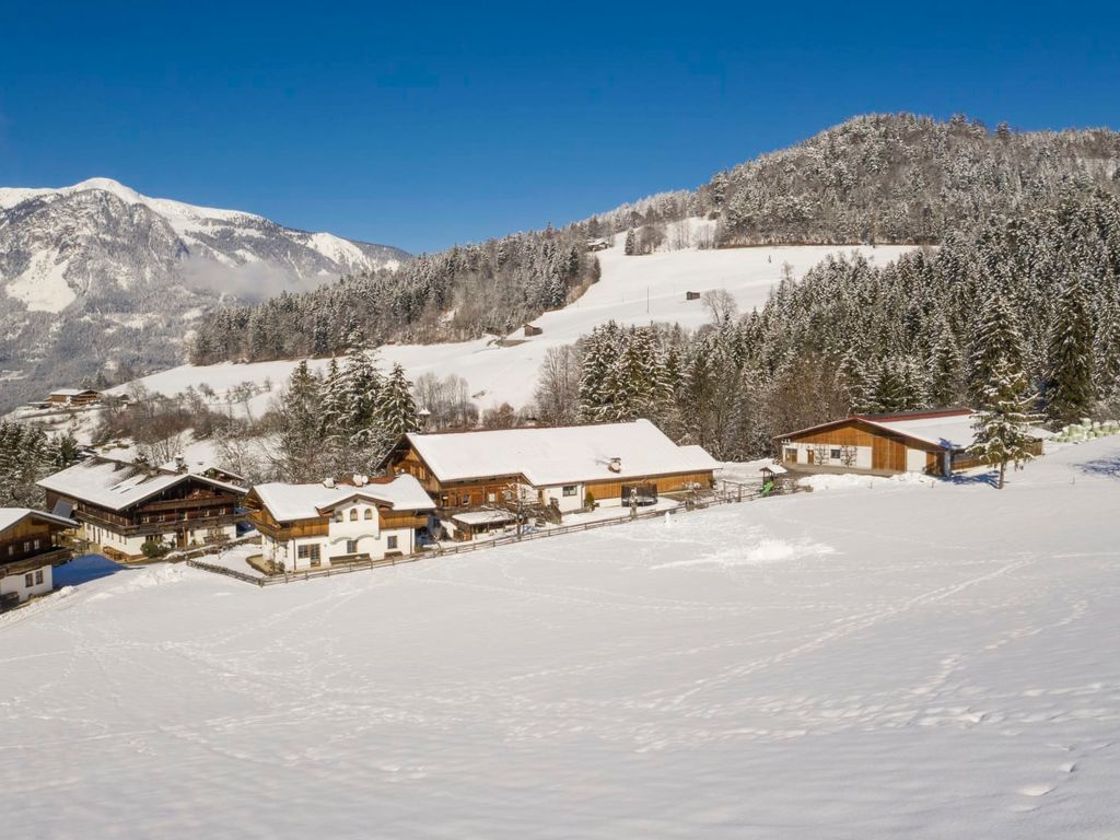 Die Außenansicht des Bauernhofs im Winter, umgeben von verschneiten Feldern, Wäldern und Bergen.