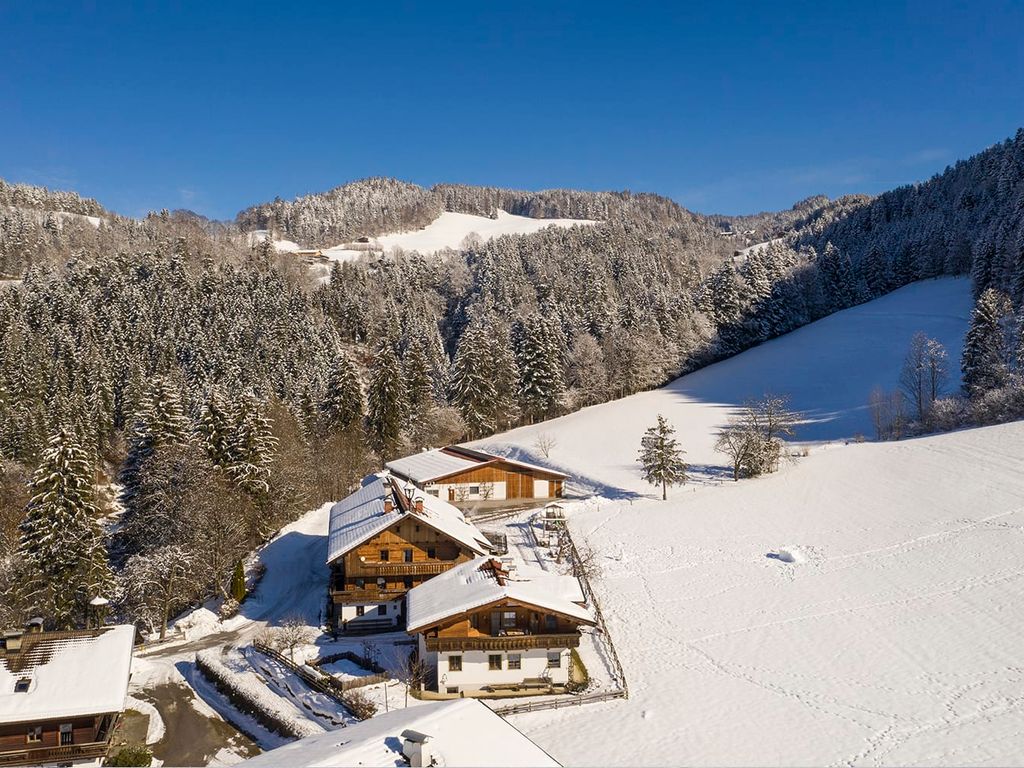 Der Bauernhof in einer schneebedeckten Winterlandschaft, umgeben von verschneiten Wäldern und Bergen.