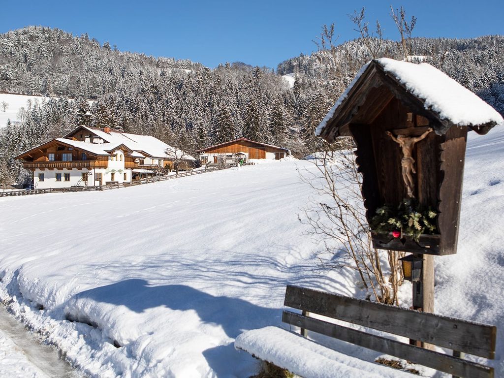 Die Außenansicht des Bauernhofs in einer verschneiten Winterlandschaft, umgeben von Bergen und einem traditionellen Wegkreuz.
