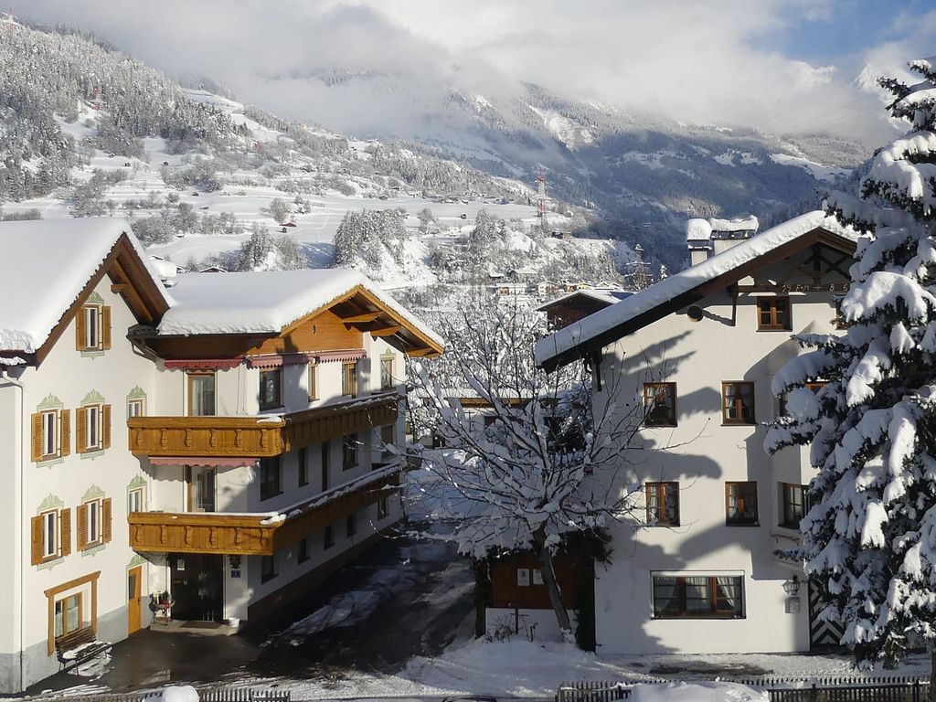 Die verschneiten Gebäude des Bauernhofs mit Balkonen und einem großen Nadelbaum vor winterlicher Bergkulisse.