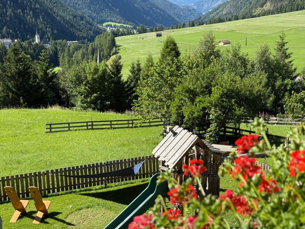 Kinderspielplatz mit Rutsche und Liegestühlen auf dem Rasen des Bauernhofs, mit Blick auf die umliegenden grünen Felder und Berge.