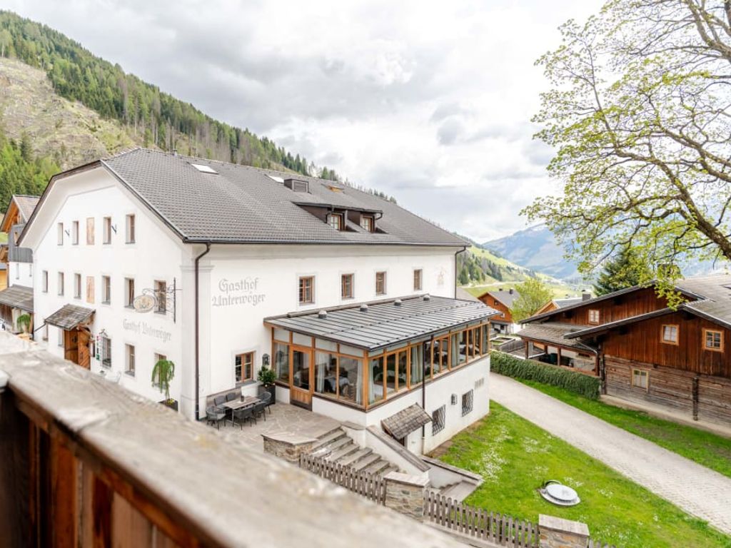 Außenansicht des Bauernhofs mit einem weißen Hauptgebäude, einem verglasten Essbereich und einer Außenterrasse, umgeben von Bergen.