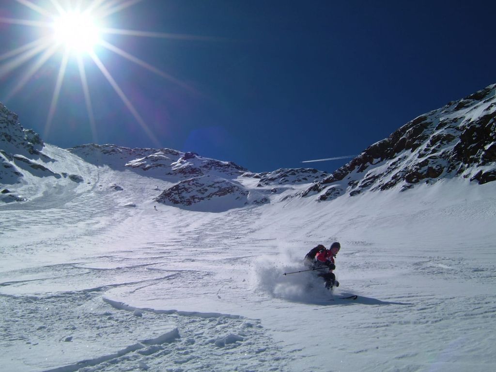 Ein Skifahrer fährt bei Sonnenschein eine verschneite Bergflanke hinunter, mit der Möglichkeit, Skiausrüstung im Trockenraum des Bauernhofs zu lagern.