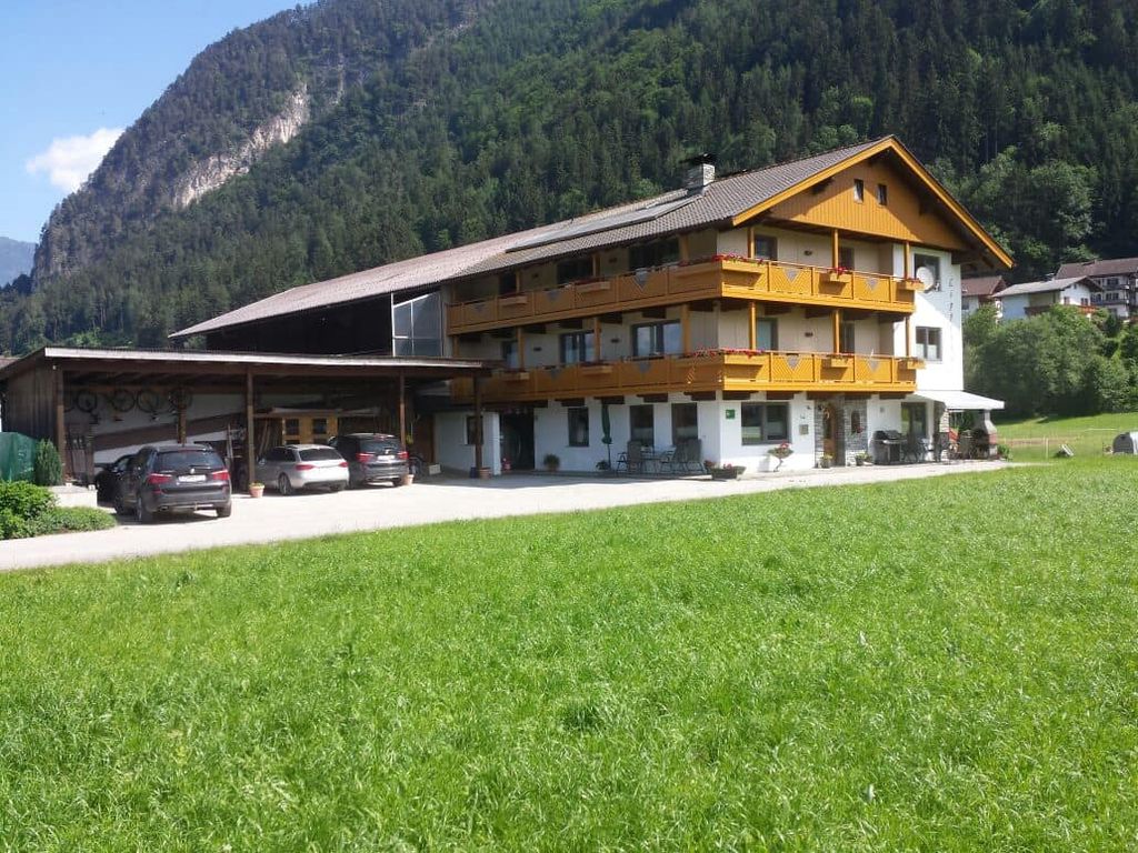Der Bauernhof mit Holzverkleidung und Balkonen, angrenzendem Unterstand für Fahrzeuge und umgeben von grünen Wiesen und Bergen.