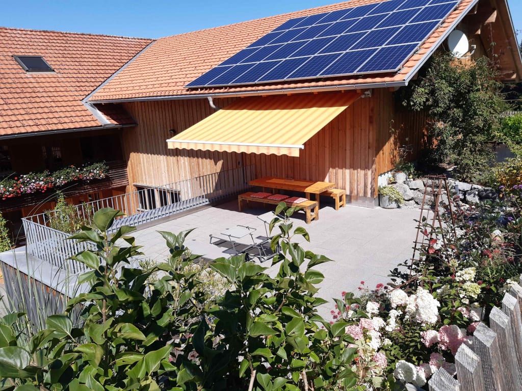 Die Sonnenterrasse am Bauernhof mit Sitzmöglichkeiten unter einer Markise und blühenden Pflanzen.