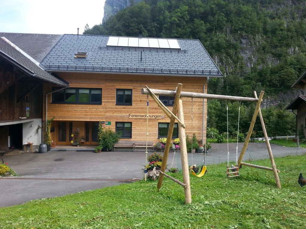 Der Bauernhof mit Holzfassade, einem Kinderspielbereich mit Schaukeln und der umliegenden Berglandschaft.