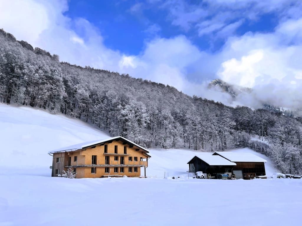 Der Ferienhof mit Holzfassade und Balkonen, eingebettet in eine schneebedeckte Winterlandschaft mit Wald und Bergen.
