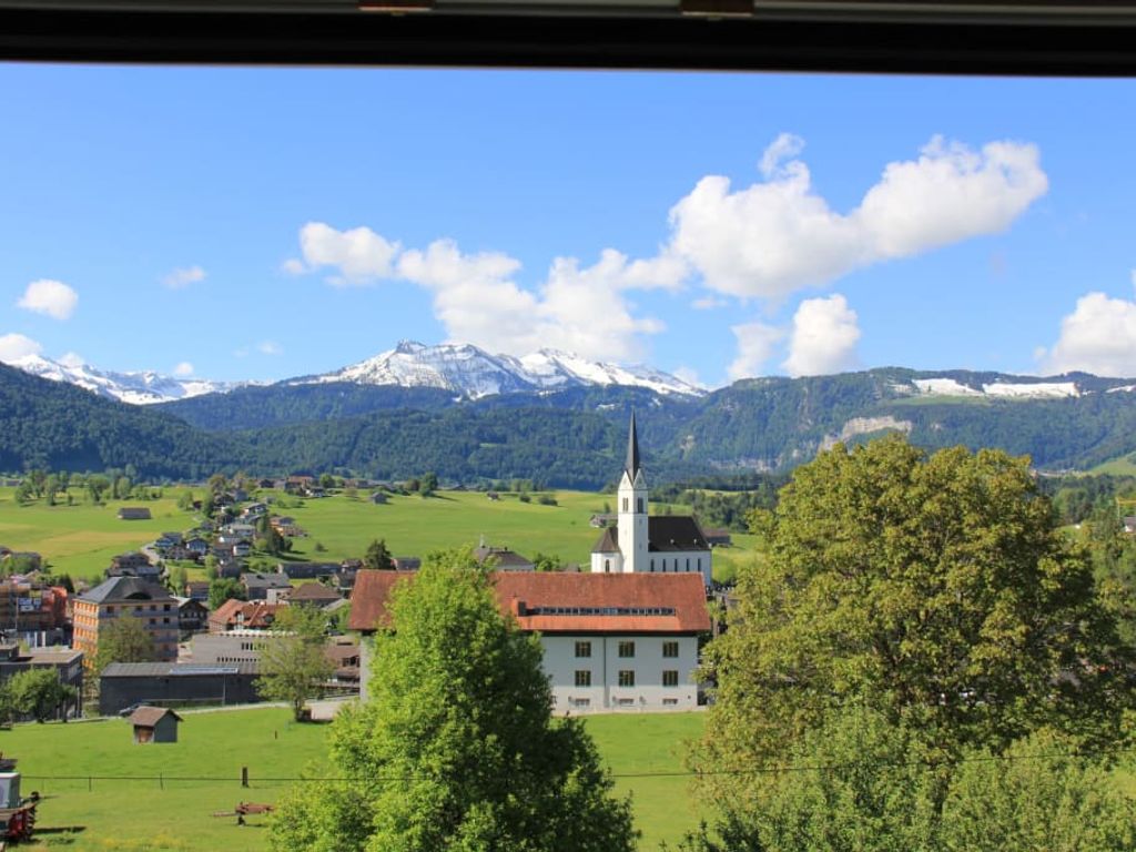 Blick aus dem Fenster auf das Dorf mit Kirche, grüne Felder und schneebedeckte Berge.