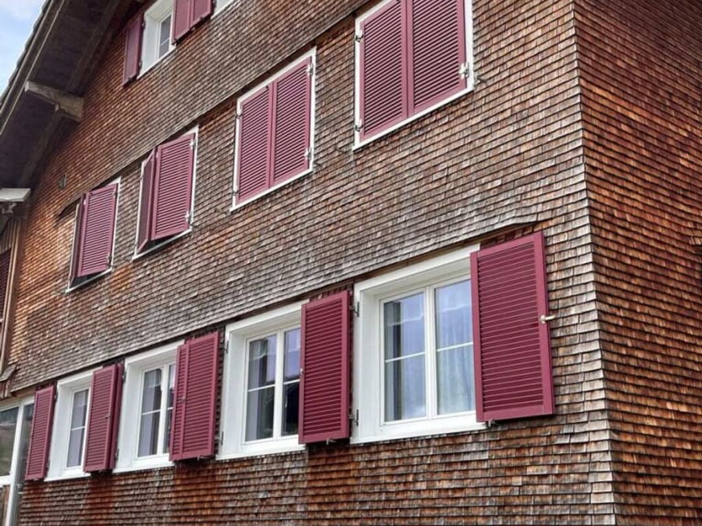Außenansicht des Bauernhofs mit Holzschindelfassade, weißen Fenstern und roten Fensterläden.