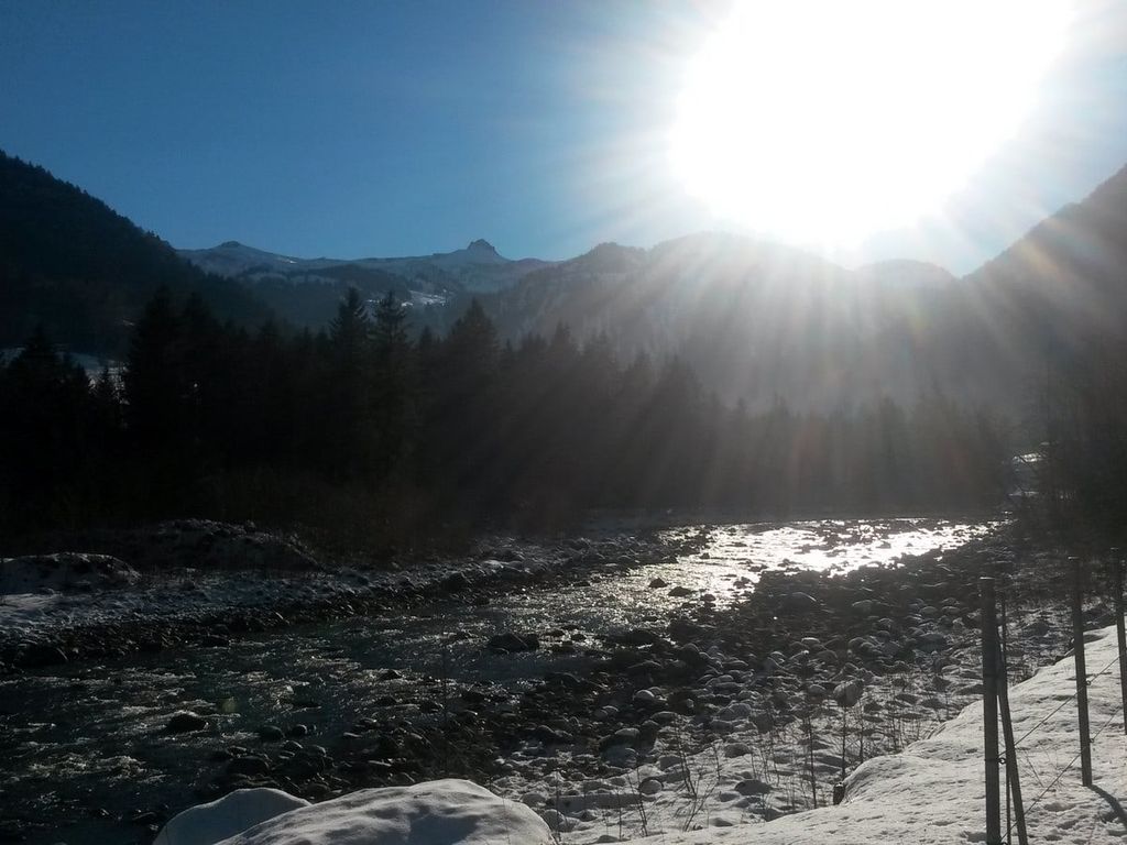 Helles Sonnenlicht glänzt auf dem Fluss in der winterlichen Berglandschaft nahe dem Bauernhof.