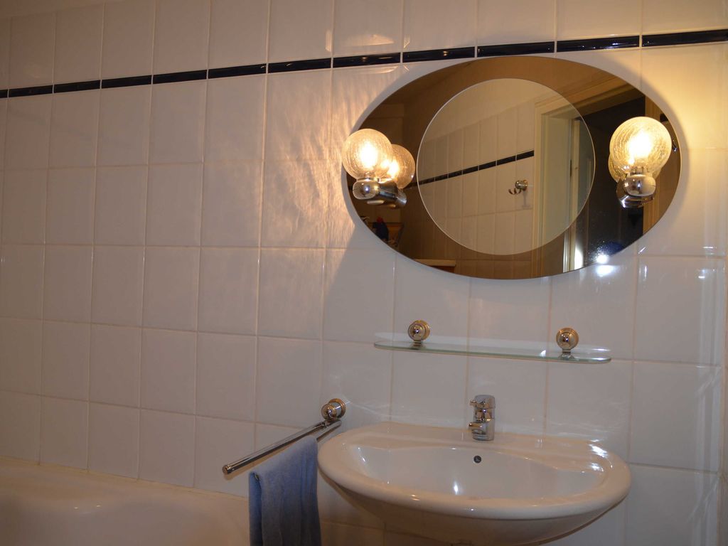 Bad mit Eckwanne Erdgeschoss. Stilvolles Badezimmer mit weiß gefliesten Wänden, modernem ovalem Spiegel, warmem Licht und Waschbecken für entspannten Wellness-Komfort.