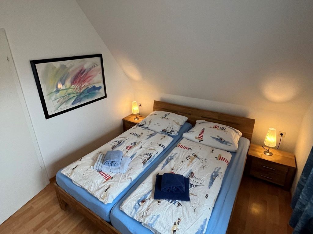 Schlafzimmer mit Doppelbett
Doppelbett aus Holz mit maritimer Bettwäsche und hellblauen Laken unter einer Dachschräge. Flankiert von zwei hölzernen Nachttischen mit brennenden Lampen, an der linken Wand hängt ein abstraktes Bild.