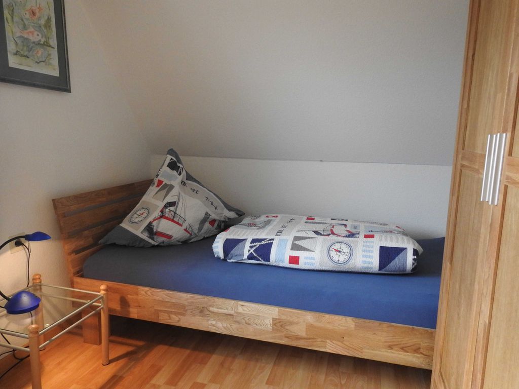 Ein gemütliches Einzelbett aus hellem Holz mit blauem Laken steht unter einer Dachschräge in einer Zimmerecke. Das Kopfkissen und die Decke haben ein maritimes Muster mit Segelbooten und Kompassen. Links neben dem Bett befindet sich ein kleiner Nachttisch aus Glas und Holz mit einer blauen Lampe, darüber hängt ein gerahmtes Bild an der Wand. Rechts im Vordergrund ist ein Teil eines hölzernen Kleiderschranks zu sehen. Der Boden besteht aus hellem Laminat.