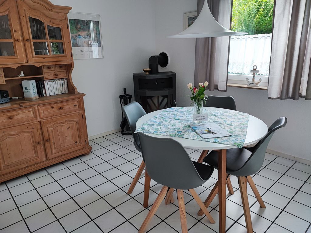 Essbereich mit einem runden weißen Tisch, vier modernen grauen Stühlen und einer Hängelampe. Im Hintergrund stehen ein rustikaler Holz-Buffetschrank, ein schwarzer Kaminofen und ein Fenster mit Vorhängen.