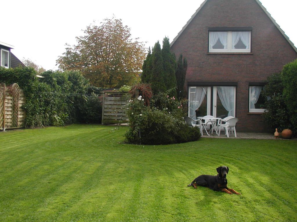 Ein großer, grüner Garten mit einer gepflegten Rasenfläche, auf der ein schwarzer Hund liegt. Im Hintergrund steht ein Einfamilienhaus aus rotem Backstein mit einer Terrasse, weißen Gartenmöbeln und dichten Hecken an den Grundstücksgrenzen.