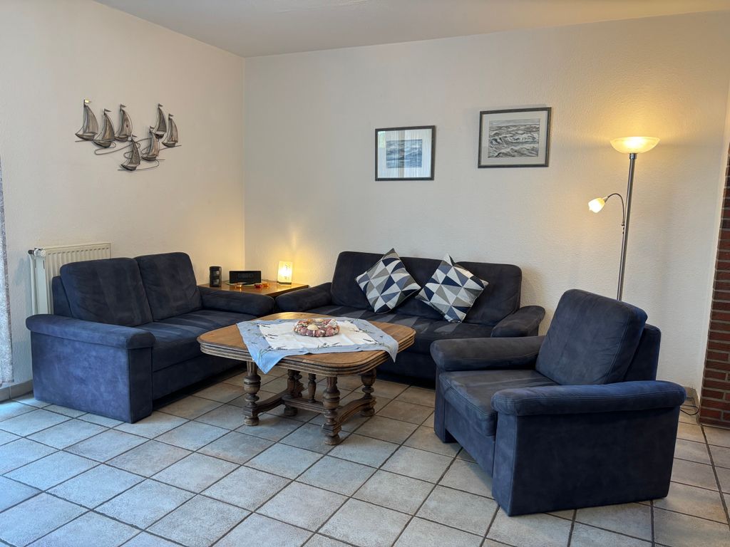 Wohnzimmer Eine gemütliche Sitzecke bestehend aus zwei dunkelblauen Sofas, einem passenden Sessel und einem rustikalen Holz-Couchtisch in der Mitte. An den weißen Wänden hängen eine Wanddekoration in Form von Segelbooten sowie zwei Bilder, während eine Stehlampe und eine Tischlampe den gefliesten Raum warm erleuchten.