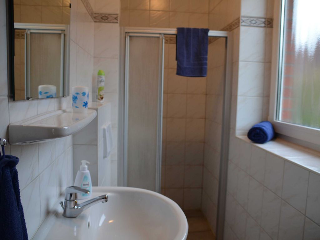 Bad im Erdgeschoss Hell gefliestes Badezimmer mit einem weißen Waschbecken und Spiegel im Vordergrund. Im Hintergrund befindet sich eine Dusche mit Schiebetür, an der ein blaues Handtuch hängt, sowie ein großes Fenster mit Milchglas.