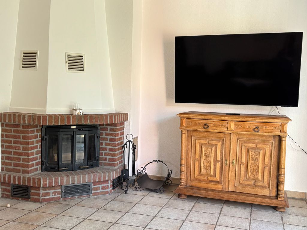 Kamin und Fernsehbereich Ein großer Flachbildfernseher hängt an einer weißen Wand über einer rustikalen Holzkommode. Links daneben befindet sich ein gemauerter Eckkamin aus roten Backsteinen mit Kaminbesteck und einer Holzschale auf dem hellen Fliesenboden.