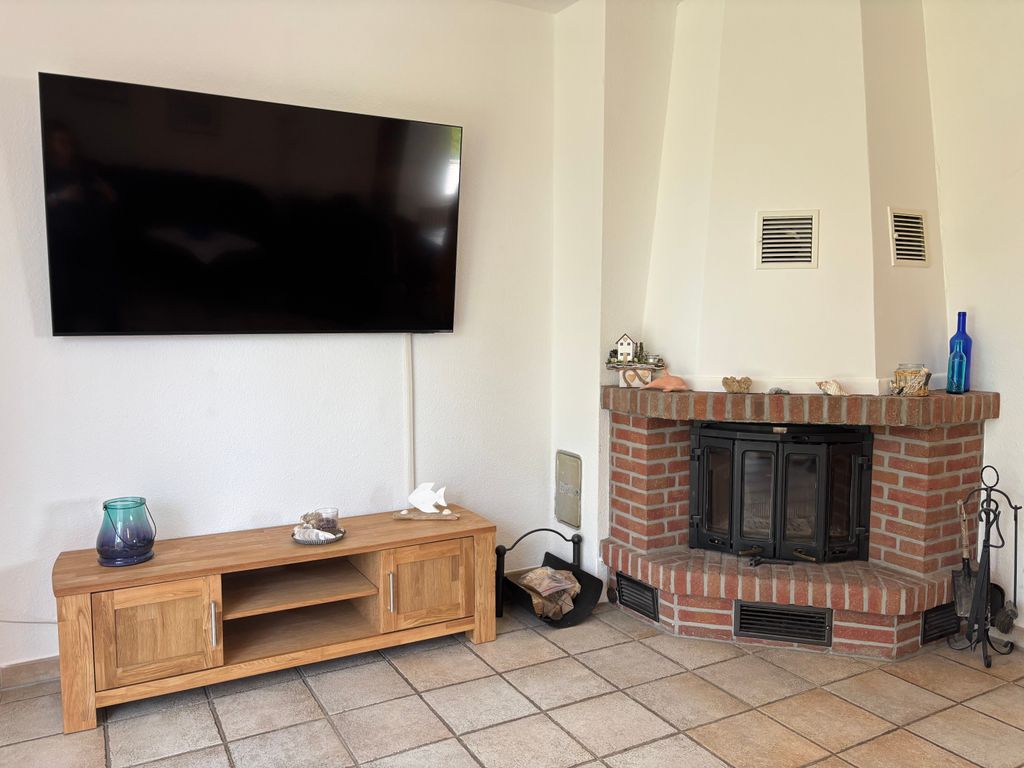 TV und Kamin Ein großer, an der weißen Wand montierter Flachbildfernseher über einem niedrigen TV-Lowboard aus hellem Holz. Rechts daneben befindet sich ein gemauerter Eckkamin aus roten Backsteinen mit Kaminbesteck und Deko-Elementen auf der Ablage, platziert auf hellem Fliesenboden.