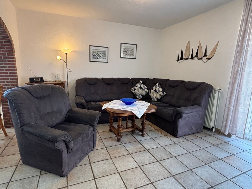 Wohnzimmer Ein gemütlicher Wohnbereich mit einer großen, dunkelgrauen Eckcouch, einem passenden Sessel im Vordergrund und einem rustikalen Holz-Couchtisch. An der weißen Wand hängen zwei Bilder und eine Holzdekoration in Form von Segelbooten, beleuchtet von einer Stehlampe in der Ecke.
