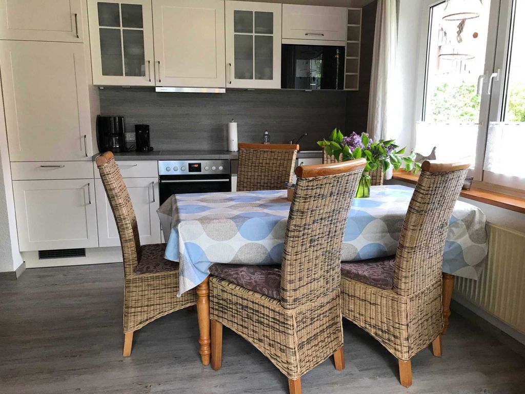 Küchenansicht Helle Küche im Ferienhaus im Landhausstil mit gemütlicher Essgruppe aus Rattanstühlen, gemusterter Tischdecke und Blick auf das Fenster.