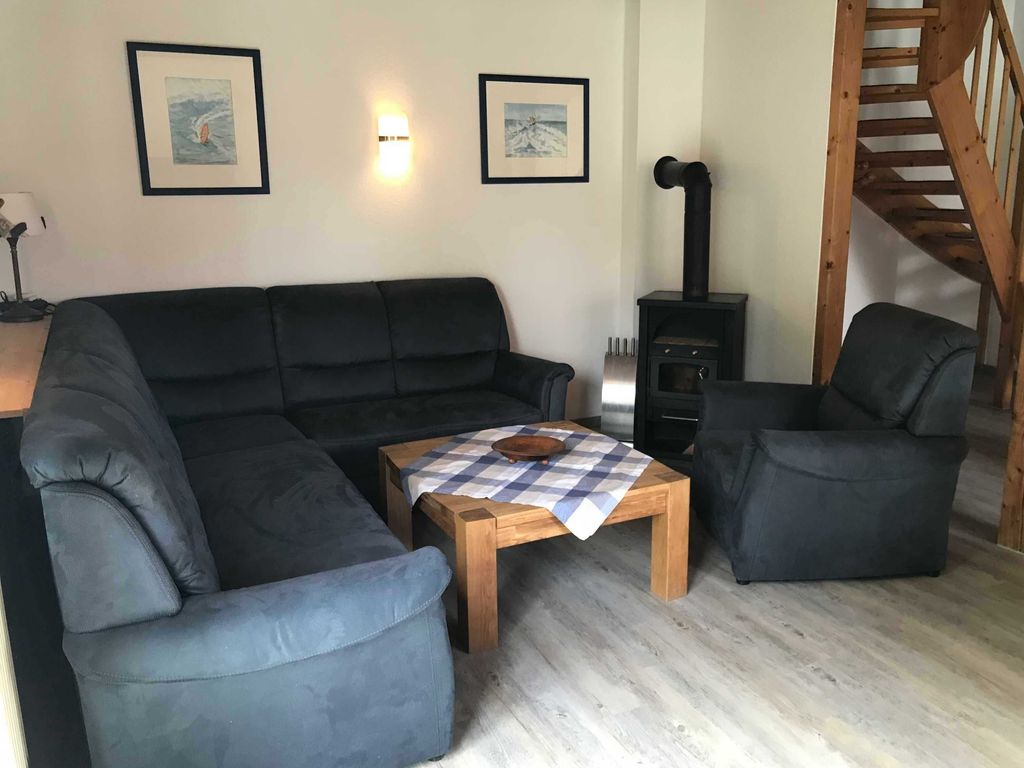 Wohnzimmer mit Kamin Gemütliches Wohnzimmer im Ferienhaus mit einer dunkelblauen Couch-Eckkombination, einem Sessel, Holz-Couchtisch und einem Kaminofen neben der Treppe.