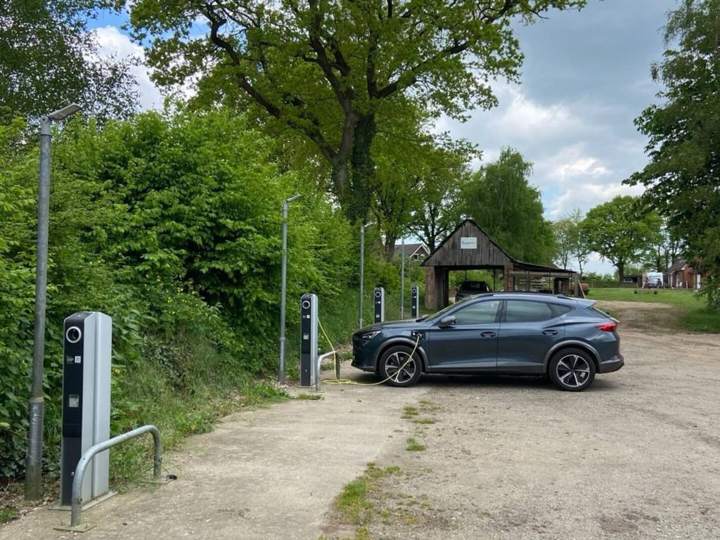 Ein Parkplatz im Freien mit mehreren modernen, silber-schwarzen E-Ladesäulen. Ein dunkelgraues Elektroauto ist an eine der Säulen angeschlossen und lädt. Der Bereich ist von hohen grünen Bäumen und Büschen umgeben, im Hintergrund ist ein hölzerner Unterstand zu sehen. Der Boden besteht aus einer Mischung aus Betonplatten und Kiesweg.