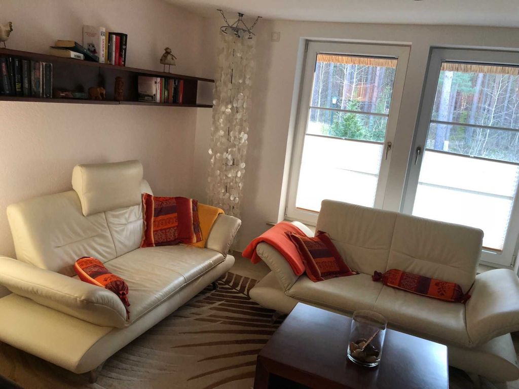 Ferienhaus Dünenwald 1 in Karlshagen auf Usedom - Das gemütliche Wohnzimmer verfügt über eine bequeme Couchgarnitur mit Tisch und lädt zum Entspannen und Verweilen ein. Die großen Fenster sorgen für viel Tageslicht und schaffen eine helle, angenehme Atmosphäre. Gleichzeitig genießen Sie einen schönen Blick ins Grüne und auf die umliegende Natur.