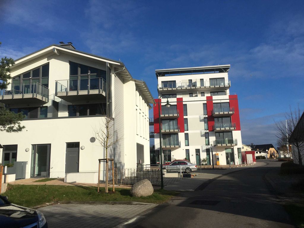 Haus Bodden-Markt Glowe von der Straße: heller Neubau mit Glasbalkonen, daneben das Haus mit den roten Fassadenfeldern